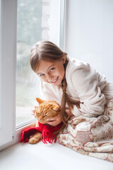 Little girl in teddy robe sitting on a window sill with pet. Lazy winter weekend with red cat at home.