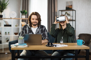 Happy young friends sitting at the table and using modern gadgets for work or study. Pretty girl wearing VR glasses and enjoying artificial reality, while focused man is typing on laptop.