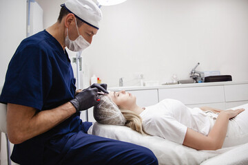 A male cosmetologist in black gloves injects botulinum toxin into the patient's forehead. A blonde...