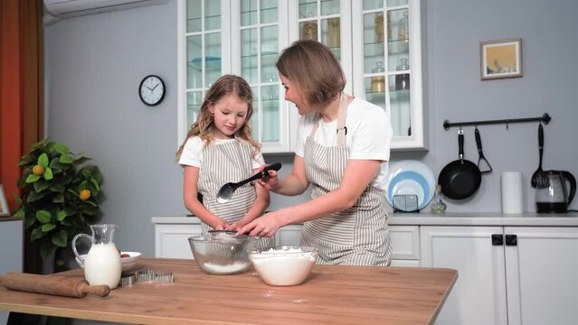 motherhood, loving mother together with her daughter repare buns and pour flour into bowl while standing at table in kitchen
