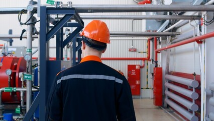 Follow the footage of a factory worker in a hard hat walking through industrial facilities. View from the back of a worker in special clothes who is walking through the plant. Factory worker.