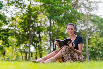 Fototapeta premium Portrait photo of the beautiful moment of a young beautiful asian female lady sitting on the grass and reading a book in an outdoor park during her summer holidays
