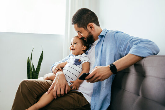 Happy Brazilian father and baby son laughing sitting on the sofa. Brazilian father holding phone with cute little boy at home.