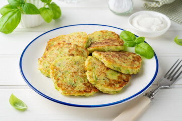 Zucchini fritters with cheese, garlic and herbs. Vegetarian zucchini pancakes, served with basil and sour cream on a white wooden background. Selective focus.