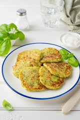 Zucchini fritters with cheese, garlic and herbs. Vegetarian zucchini pancakes, served with basil and sour cream on a white wooden background. Selective focus.
