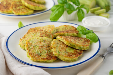 Zucchini fritters with cheese, garlic and herbs. Vegetarian zucchini pancakes, served with basil and sour cream on a white wooden background. Selective focus.