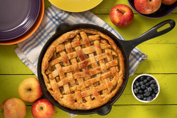 Apple pie in a cast iron skillet on a green coor table surrounded by apples.