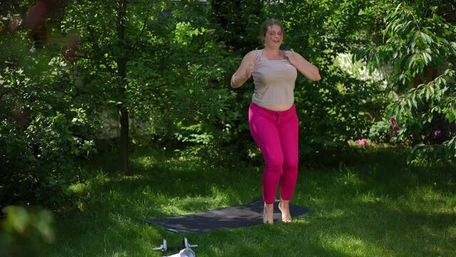 Barefoot plus-size woman running in circles in summer garden smiling. Wide shot portrait of motivated overweight Caucasian sportswoman working out outdoors having fun. Sport and happiness