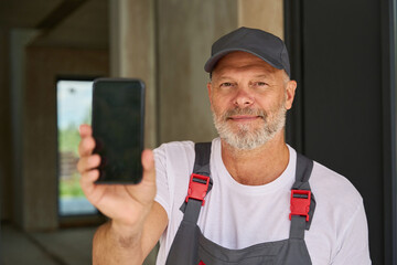 Adult builder in overalls and baseball cap shows phone