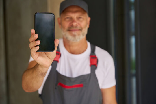 Smiling builder wearing cap and overalls shows mobile phone