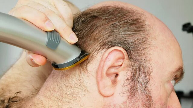 Man shaves his head. An overgrown man with a bald patch clumsily cuts his brown gray hair on his head with an electric clipper. Self shaving head with a trimmer. Amateur haircut in the bathroom