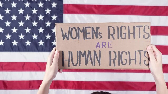 Woman from the back holding a sign Womens Rights Are Human Rights US flag on background. Protest against anti abortion law. Women's strike. Womens rights freedom.