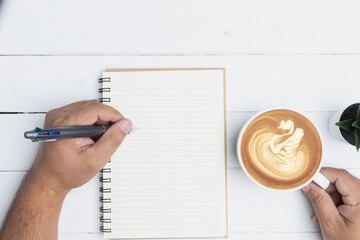 Top view with copy space, flat lay. White office desk table with a blank notebook, a pen, and a cup of coffee on old wooden background. Hand man writing record on a notebook in the office. - obrazy, fototapety, plakaty