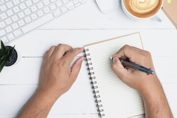 Hand man writing record on a notebook in the office. White office desk table with a blank notebook, a pen, a keyboard, and a cup of coffee on old wooden background. Top view with copy space, flat lay. - obrazy, fototapety, plakaty