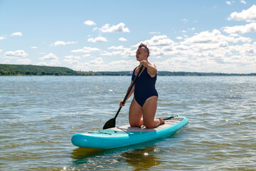 a woman in a closed swimsuit with kneeling on a SUP board with a paddle floats on the water against the blue sky.