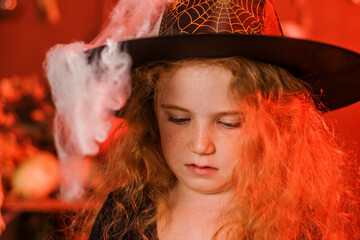 Portrait of a cute red-haired girl in a black witch costume and hat with a cobweb in Halloween decorations.Halloween concept.