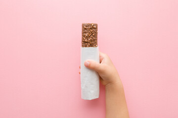 Baby girl hand holding dark brown chocolate bar with nuts on light pink table background. Pastel color. Sweet snack in opened white pack. Closeup. Top down view.