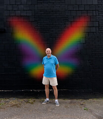 A mature gay man stands outside, in front of a black brick wall with rainbow colored faerie /...