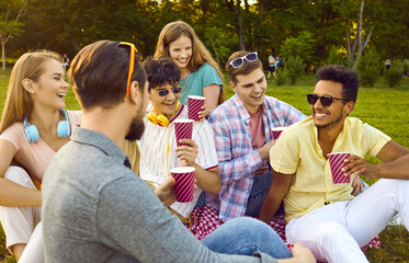 Group of friends gather together in summer park and talk on picnic with paper cups of coffee or tea. Cheerful multiracial and young people having fun, laughing, talking and drinking beverages outdoors