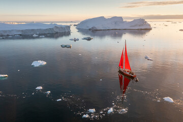 Sail boat with red sails cruising among ice bergs during dusk in front of a full moon. Disko Bay,...