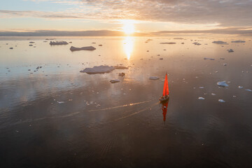 Sail boat with red sails cruising among ice bergs during dusk in front of a full moon. Disko Bay,...