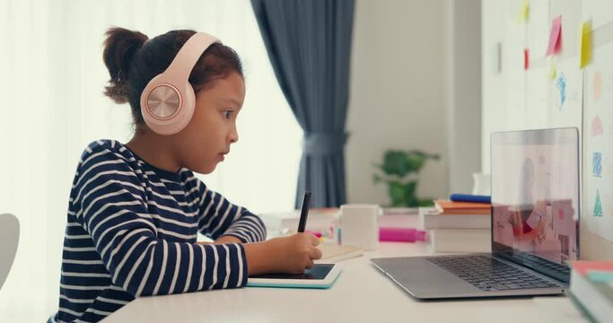 Asian toddler girl with sweater wear headphone sit front of desk with notepad use magic pen focus on writing do homework, finger counting number math online learning course from laptop at home.