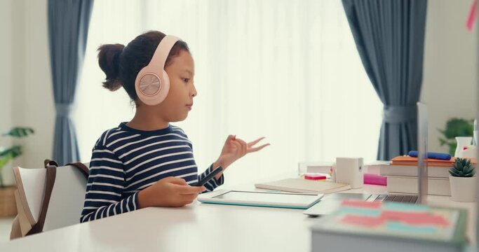 Asian toddler girl with sweater wear headphone sit front of desk with notepad use magic pen focus on writing do homework, finger counting number math online learning course from laptop at home.