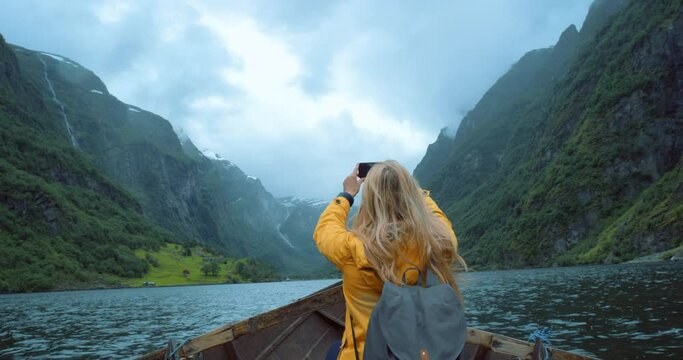 Female travel researcher taking phone photos of green mountain views, sitting alone in a row boat. Back view of woman doing ecology research outside. Traveler takes a picture for online content