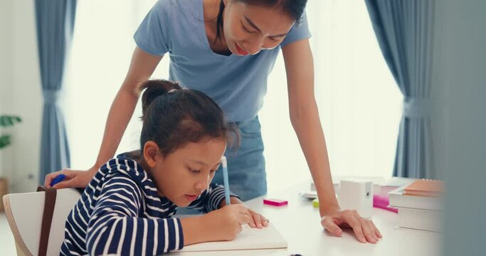 Asian toddler girl with sweater sit in front of desk with notepad use pencil focus on write notebook do math homework with her mother or tutor on weekend at home. Distance online learning concept.