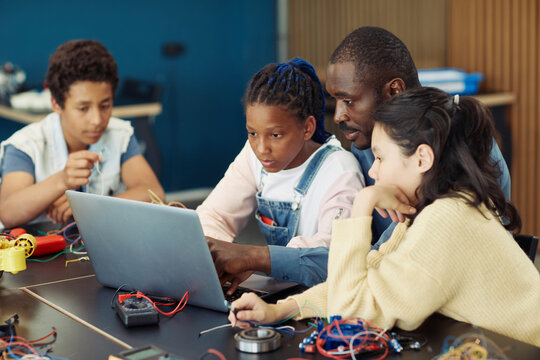 Portrait of black teenage girl using laptop in school during engineering class with male teacher and diverse group of children