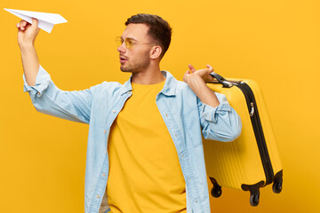 Smiling joyful handsome man in blue shirt throws paper airplane ready for vacation with suitcase posing isolated on orange yellow studio background. Copy space Banner Mockup. Trip journeys concept