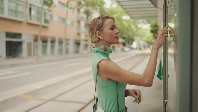 Beautiful woman with short blond hair in casual clothes stands at tram stop, looks at the public transport timetable on the information board