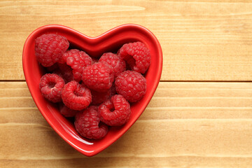 Fototapeta na wymiar Raspberries lie in a heart-shaped ceramic bowl, which stands on a wooden table. View from above