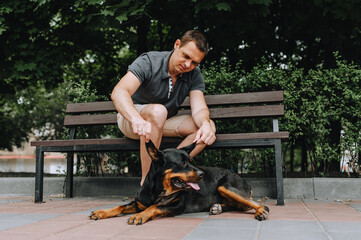 A man sits on a park bench and caresses his beloved Rottweiler dog. Photograph of man and animal.