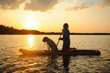 A middle-aged man with a dog paddles on the lake water on a SUP board at sunset. Silhouette of a person against the backdrop of beautiful calm nature. - obrazy, fototapety, plakaty