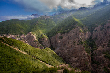 Naklejka premium Mountain Peaks Trapping Light entangled Smoky Clouds Letting few Sunrays shine on The Trees Covering The Cliffs Creating A Breathtaking Landscape Scenic View