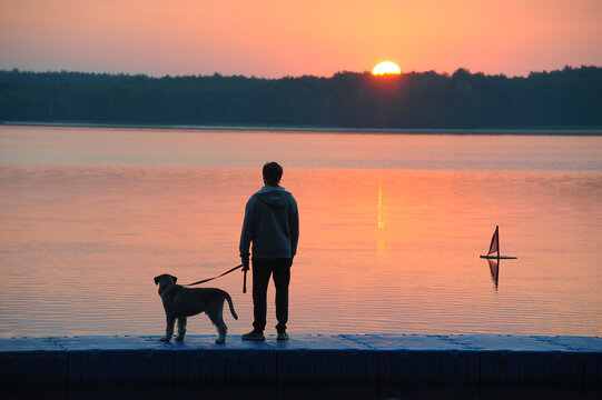 Silhouette of a middle-aged man with a dog on the shore of the lake at dawn.