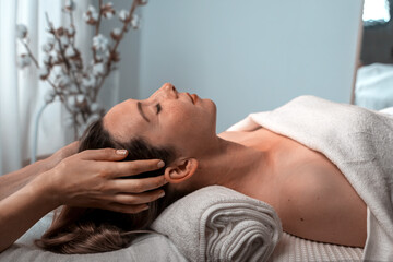 SPA procedure in a massage room with the help of hot stones, one gentel hand of a woman masseur on the female patient's face, cotton twigs on the background