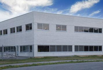 Exterior corner of nondescript two story white brick office or factory building during daytime, chain link fence, picnic table, nobody