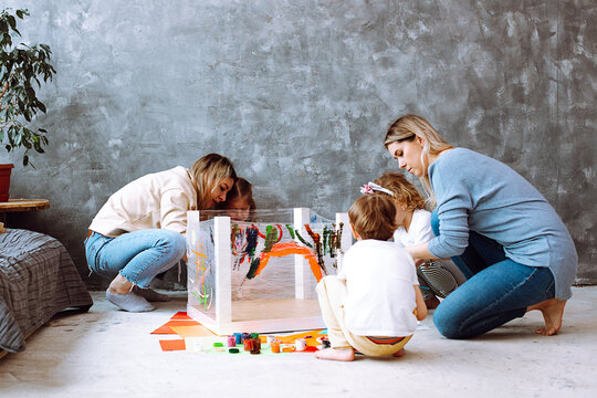 Attentive little girls sitting on haunches on art lesson, painting with mothers teachers with brushes on stretch film.