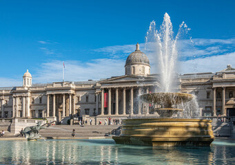 London, UK- July 4, 2022: Trafalgar Square. Wide view on Full blowing water Jellycoe fountain with National Gallery building in back under blue cloudscape. - obrazy, fototapety, plakaty