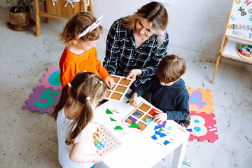 Top view of group of preschoolers. Young blond woman helping pupils to play board games, collecting...