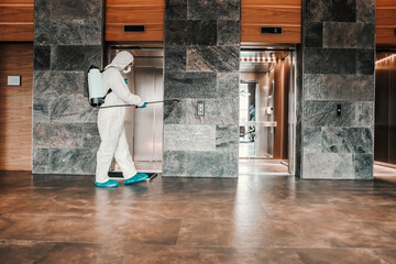 Fight against COVID19. A man in a white protective suit with protection disinfects lobby area and automatic elevator door. Coronavirus concept, new normal