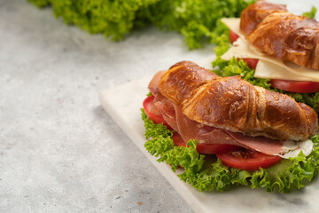 two lye croissant sandwiches with iberian ham, tomato slices, lettuce and cheese on marble board on grey background