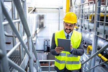 Factory manager in elegant suit and hardhat checking results at tablet computer in production plant interior.