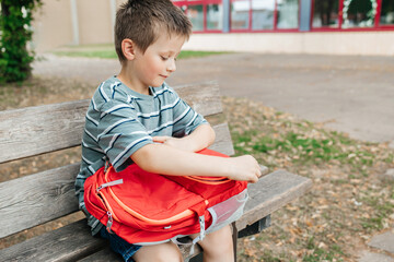 The boy sits on a bench in the school yard and takes out a lunch box from a school backpack....