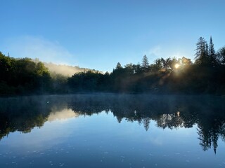Paysage calme d'une forêt et de l'eau d'une rivière à l'aube. Reflet d'une montagne sur un lac au petit matin. Lever de soleil avec brume sur l'eau. Rayons de soleil au travers le brouillard.