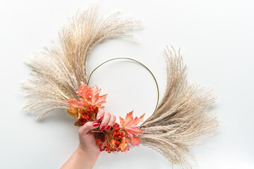Floral wreath from dry pampas grass and Autumn leaves. Hands in sweater with manicured nails tie...