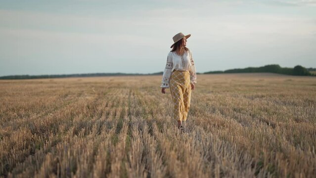 Stylish ethnic woman walking in wheat field after harvesting. Attractive lady in embroidery vyshyvanka blouse and straw hat. Ukraine, independence, freedom, patriot symbol