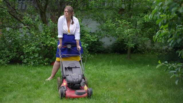 Wide shot tired woman standing with lawn mower in summer garden looking away. Portrait of Caucasian plus-size gardener with equipment outdoors on backyard. Slow motion